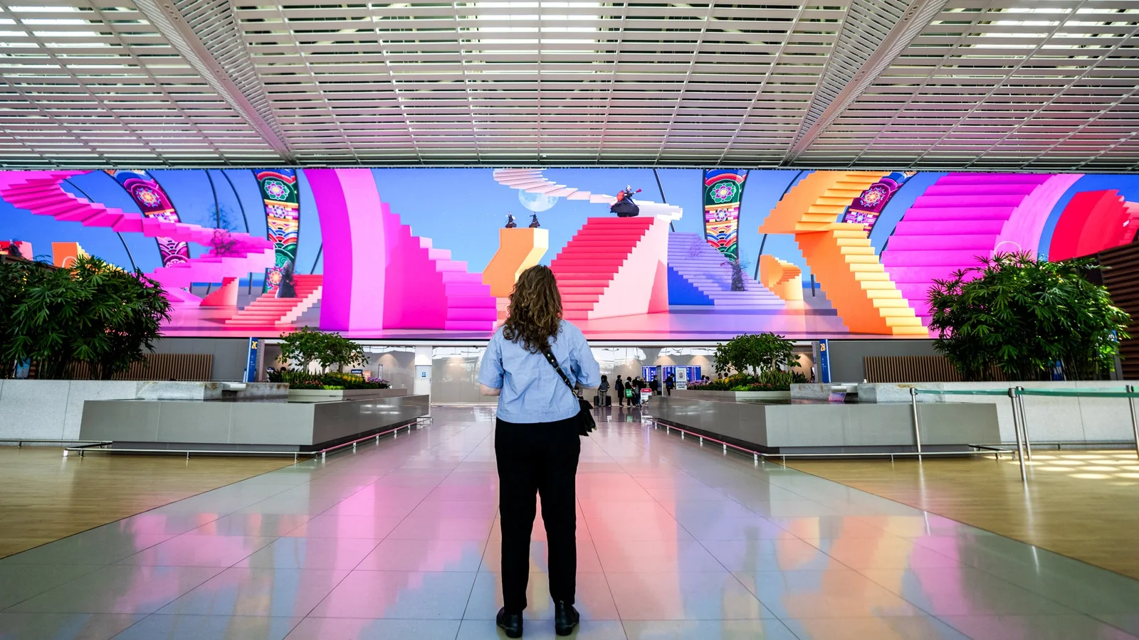 Seoul Incheon Airport Terminal 2 16K LED megacanvas by Moment Factory — a single traveller stands centred in front of a vast curved display showing geometric pink stairs as part of a Korean cultural content capsule